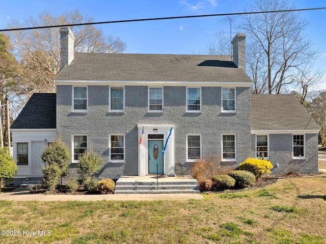 colonial house with roof with shingles, a chimney, a front lawn, and brick siding