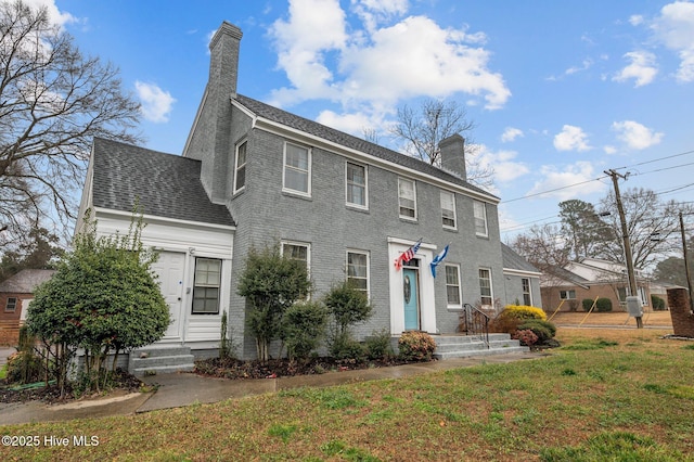 colonial inspired home featuring brick siding, a chimney, a shingled roof, entry steps, and a front yard