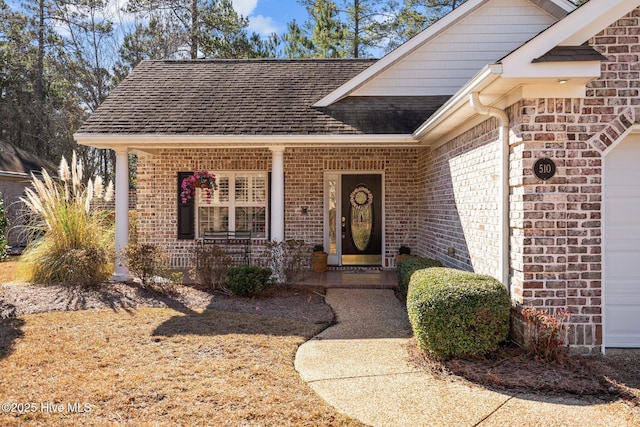 view of exterior entry with a porch, brick siding, a garage, and roof with shingles