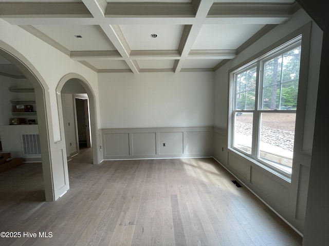 empty room with arched walkways, beam ceiling, light wood-style flooring, coffered ceiling, and a wainscoted wall
