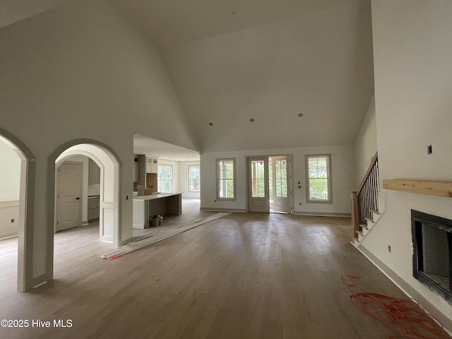 unfurnished living room with high vaulted ceiling, light wood finished floors, baseboards, arched walkways, and stairway