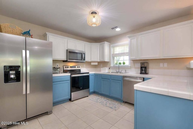 kitchen with tile countertops, blue cabinets, a sink, appliances with stainless steel finishes, and decorative light fixtures