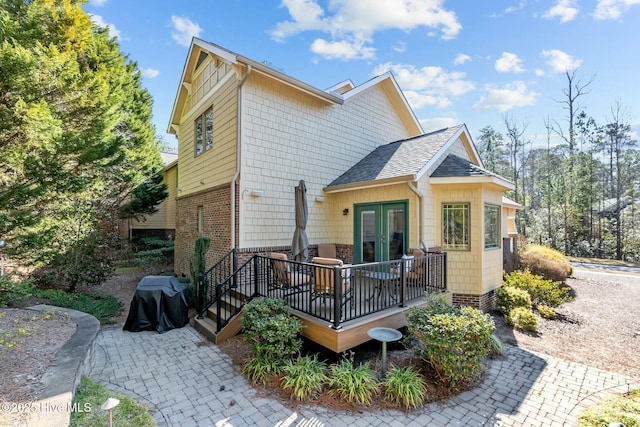 back of house with brick siding, a wooden deck, roof with shingles, and french doors