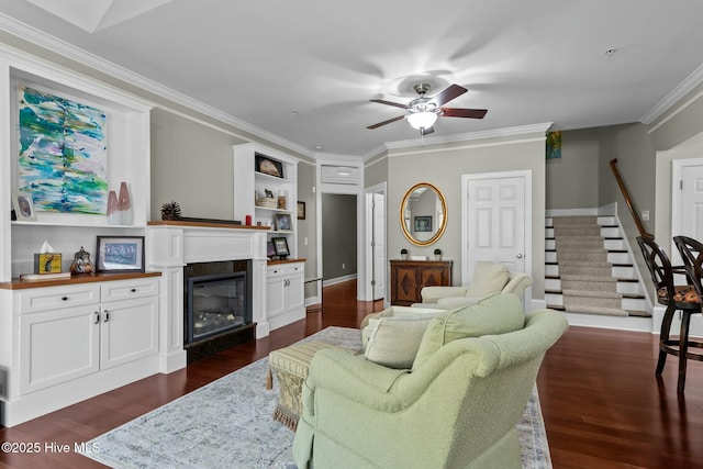 living room featuring dark wood-style flooring, crown molding, stairway, a ceiling fan, and a glass covered fireplace