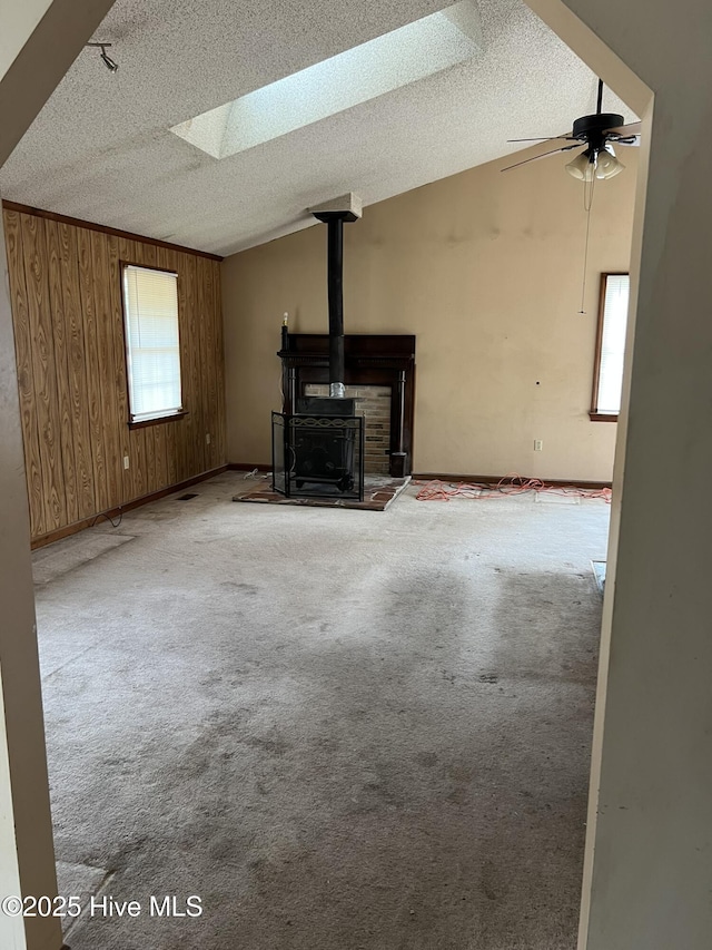 unfurnished living room with vaulted ceiling with skylight, a wood stove, carpet, and wooden walls