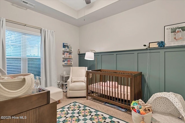 bedroom featuring a decorative wall, visible vents, wainscoting, a nursery area, and carpet