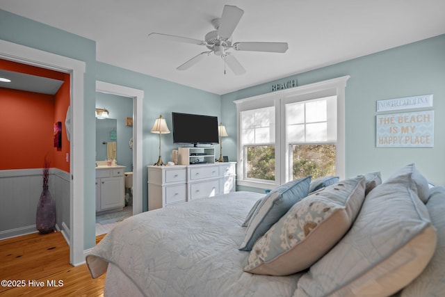 bedroom with a wainscoted wall, ceiling fan, light wood-style flooring, and ensuite bath