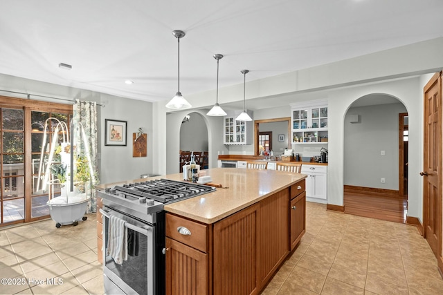 kitchen with brown cabinetry, arched walkways, stainless steel gas stove, and hanging light fixtures