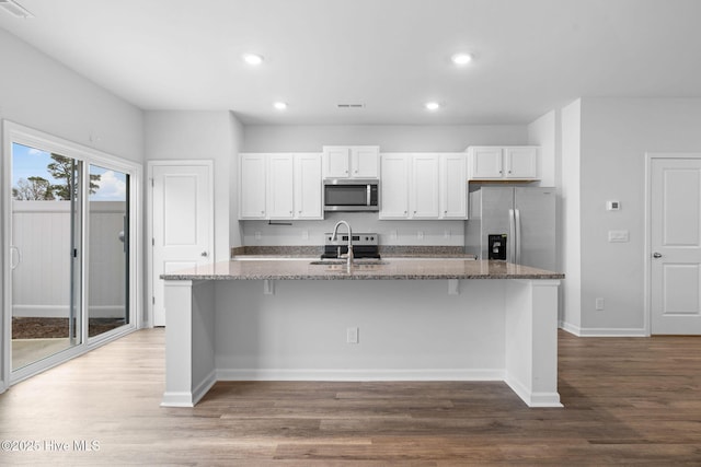 kitchen with stainless steel appliances, wood finished floors, white cabinets, dark stone counters, and an island with sink