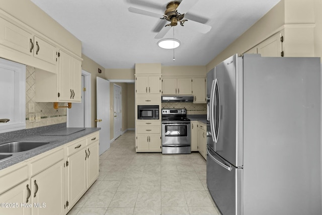 kitchen with under cabinet range hood, a sink, appliances with stainless steel finishes, cream cabinetry, and dark countertops