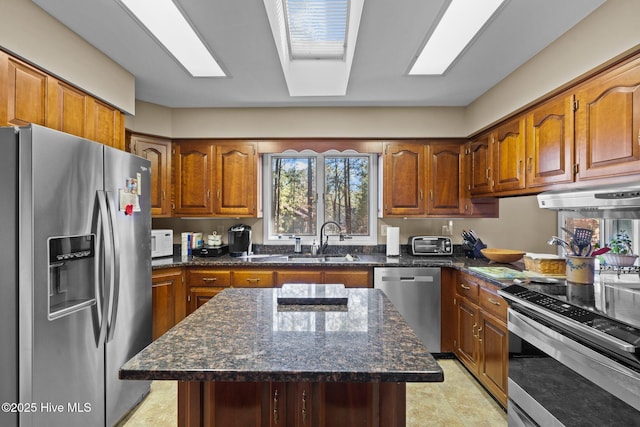 kitchen with a skylight, brown cabinets, appliances with stainless steel finishes, a sink, and under cabinet range hood