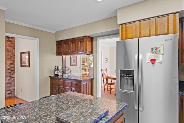 kitchen featuring baseboards, ornamental molding, stainless steel refrigerator with ice dispenser, brown cabinets, and dark stone countertops