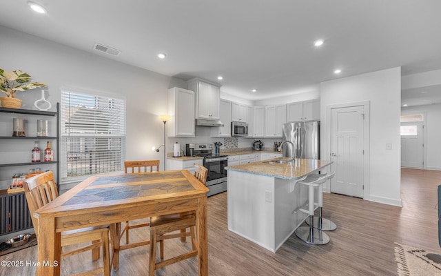 kitchen with appliances with stainless steel finishes, light hardwood / wood-style flooring, a center island with sink, light stone counters, and white cabinets