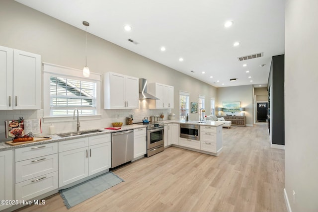 kitchen featuring stainless steel appliances, a sink, white cabinets, open floor plan, and wall chimney exhaust hood