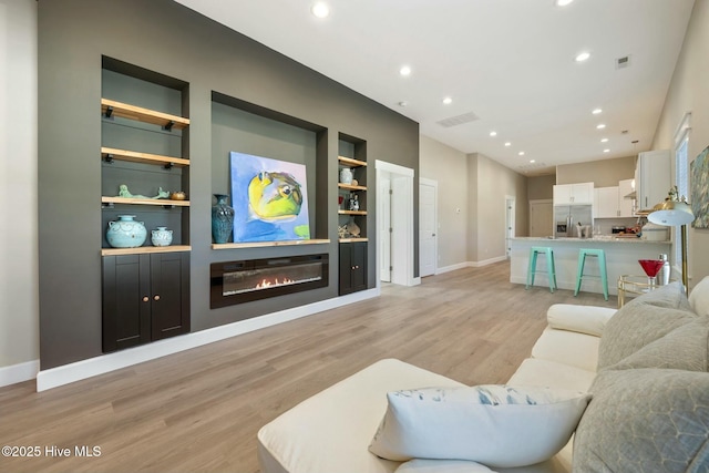 living room featuring built in shelves, a glass covered fireplace, light wood finished floors, and recessed lighting