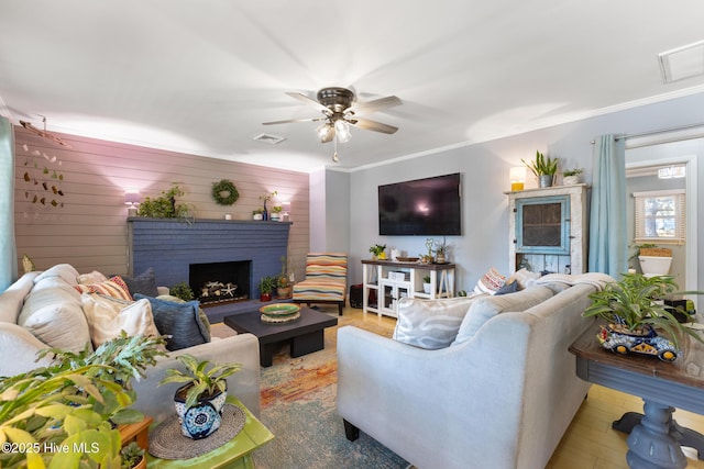 living room featuring ceiling fan, wood finished floors, visible vents, ornamental molding, and a brick fireplace