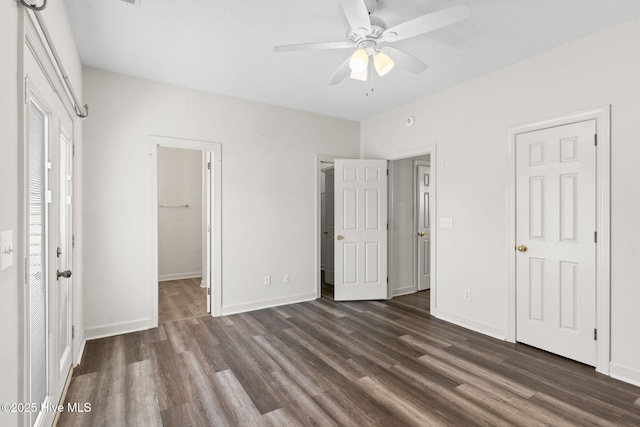 unfurnished bedroom with dark wood finished floors, a walk in closet, a closet, baseboards, and ceiling fan