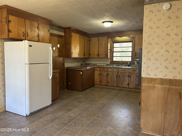 kitchen with freestanding refrigerator, brown cabinets, a sink, and wallpapered walls