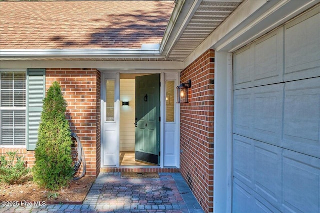 doorway to property with a shingled roof, brick siding, and a garage