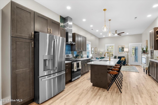 kitchen with a sink, a kitchen breakfast bar, light countertops, appliances with stainless steel finishes, and wall chimney range hood