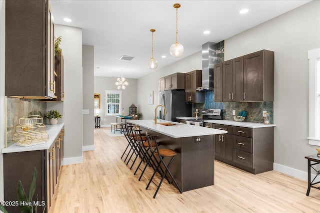 kitchen with stainless steel appliances, a breakfast bar, light countertops, wall chimney range hood, and a center island with sink