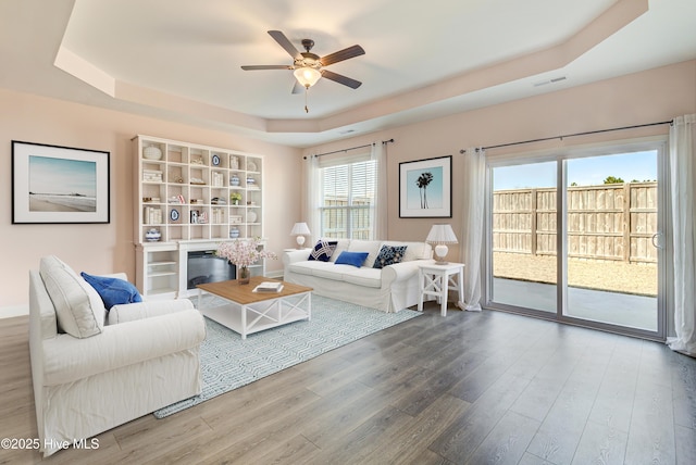 living area featuring visible vents, a ceiling fan, a glass covered fireplace, wood finished floors, and a tray ceiling