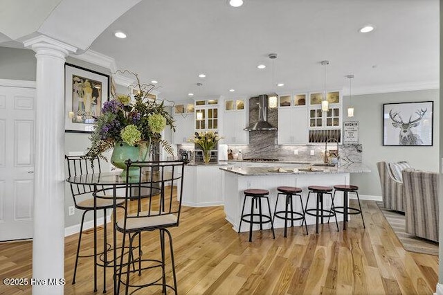 kitchen with wall chimney exhaust hood, hanging light fixtures, glass insert cabinets, white cabinets, and ornate columns