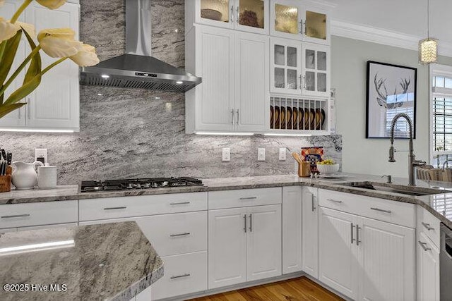 kitchen featuring a sink, white cabinetry, hanging light fixtures, wall chimney exhaust hood, and glass insert cabinets