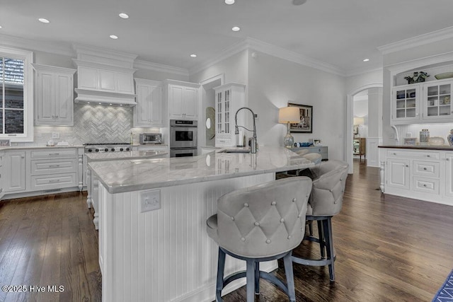 kitchen with arched walkways, white cabinets, glass insert cabinets, a large island with sink, and a sink