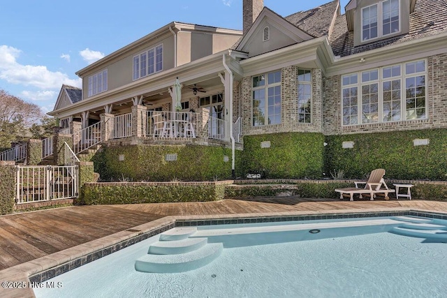 rear view of property with brick siding, a ceiling fan, a wooden deck, an outdoor pool, and a chimney