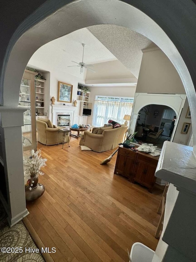 living area featuring arched walkways, a tray ceiling, light wood-style flooring, a ceiling fan, and a glass covered fireplace