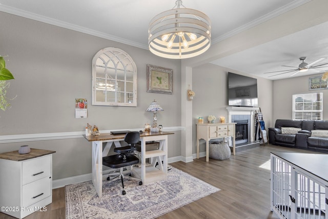 home office featuring a fireplace with raised hearth, ceiling fan with notable chandelier, wood finished floors, baseboards, and crown molding