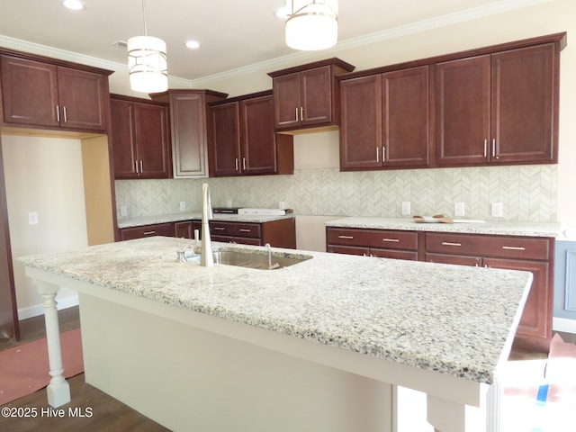 kitchen with decorative light fixtures, ornamental molding, a sink, an island with sink, and light stone countertops