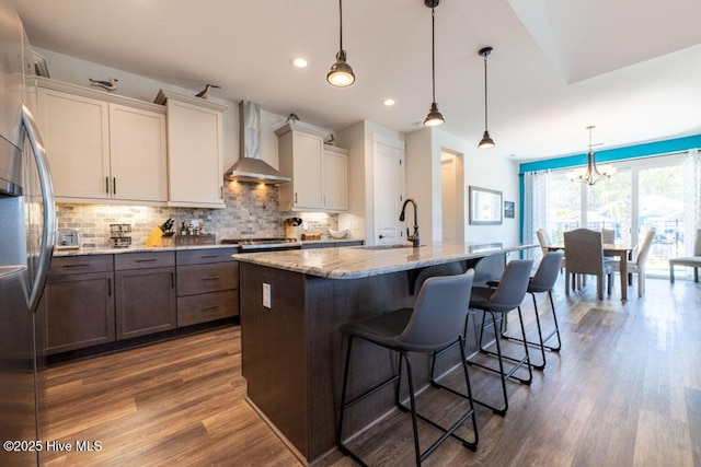 kitchen with stainless steel fridge, hanging light fixtures, a kitchen island with sink, wall chimney range hood, and a sink