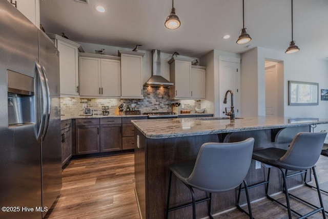 kitchen featuring hanging light fixtures, wall chimney range hood, a center island with sink, and stainless steel refrigerator with ice dispenser