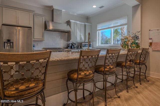 kitchen with wall chimney exhaust hood, light stone countertops, stainless steel appliances, light wood-type flooring, and backsplash