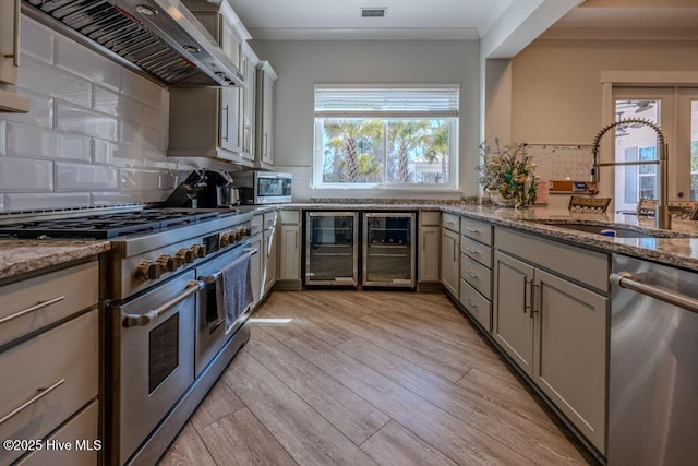 kitchen with wine cooler, stone counters, stainless steel appliances, a sink, and wall chimney range hood