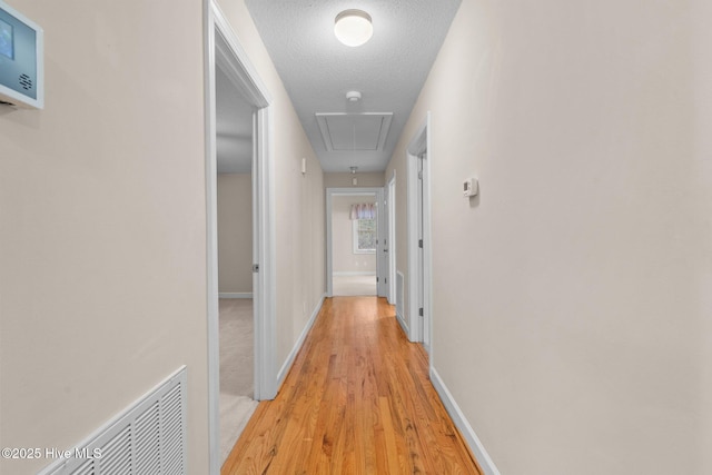 hallway featuring a textured ceiling, visible vents, baseboards, light wood finished floors, and attic access