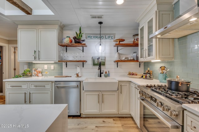 kitchen featuring open shelves, glass insert cabinets, appliances with stainless steel finishes, a sink, and wall chimney exhaust hood