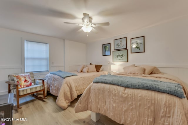 bedroom featuring light wood-type flooring, a wainscoted wall, a decorative wall, and a ceiling fan