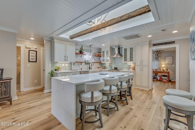 kitchen featuring glass insert cabinets, a center island, light countertops, wall chimney range hood, and white cabinetry