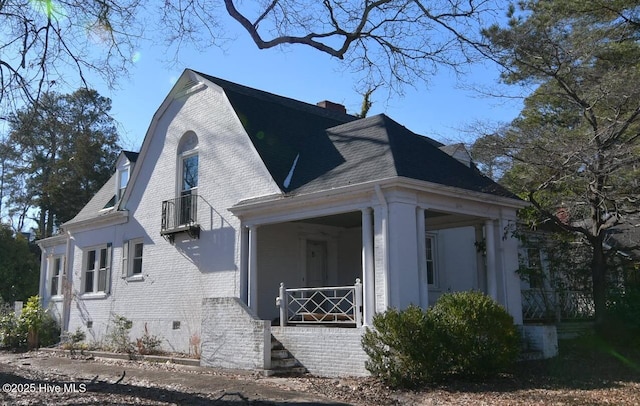 view of side of property with covered porch, brick siding, a shingled roof, a gambrel roof, and crawl space