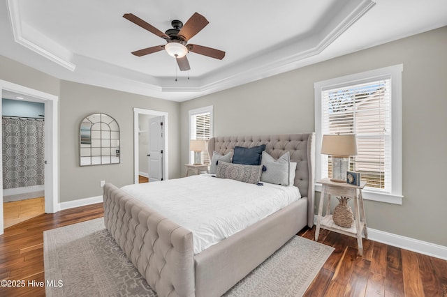 bedroom with a tray ceiling, a ceiling fan, ensuite bath, baseboards, and hardwood / wood-style flooring