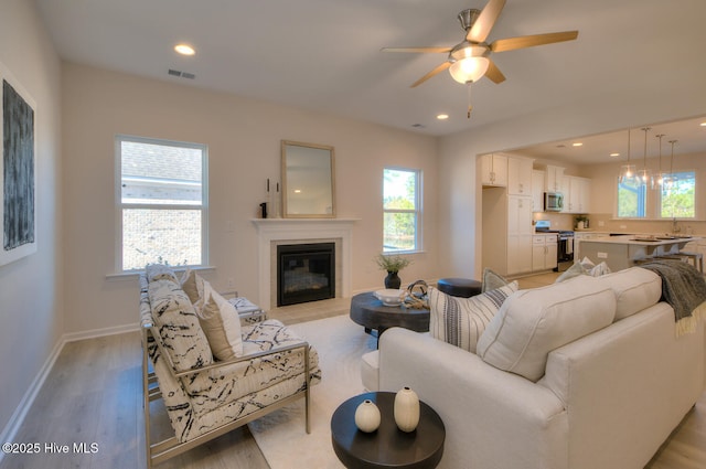 living area with recessed lighting, light wood-style flooring, a ceiling fan, a glass covered fireplace, and baseboards