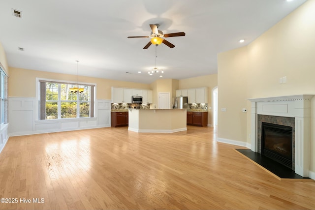 unfurnished living room with a decorative wall, ceiling fan with notable chandelier, a fireplace with flush hearth, visible vents, and light wood-style floors