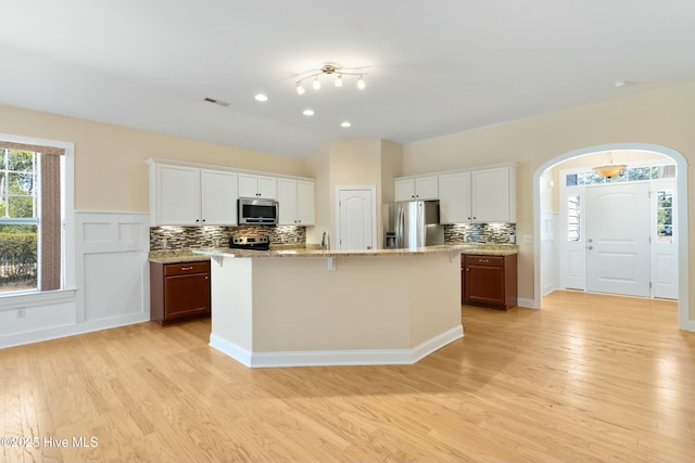 kitchen featuring arched walkways, stainless steel appliances, a center island with sink, and white cabinetry