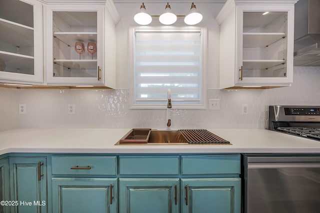 kitchen with stainless steel appliances, a sink, light countertops, wall chimney exhaust hood, and glass insert cabinets