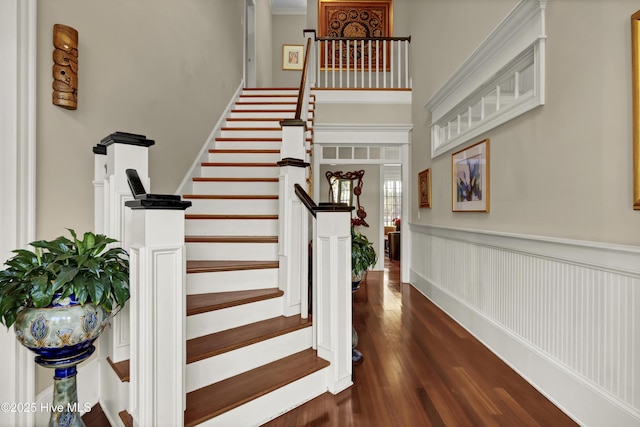 staircase with a high ceiling, wood finished floors, and wainscoting