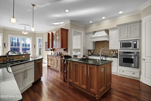 kitchen with stainless steel appliances, custom exhaust hood, washing machine and dryer, a sink, and a center island with sink