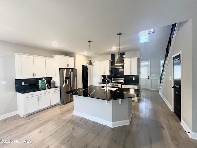 kitchen featuring white cabinets, appliances with stainless steel finishes, a center island with sink, wall chimney exhaust hood, and decorative light fixtures